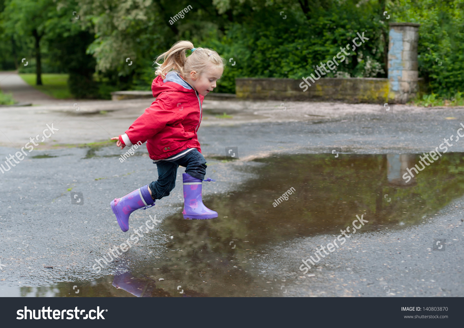 非常可爱的小女孩在红夹克,牛仔裤和橡胶靴跳过水坑在雨天-人物,公园